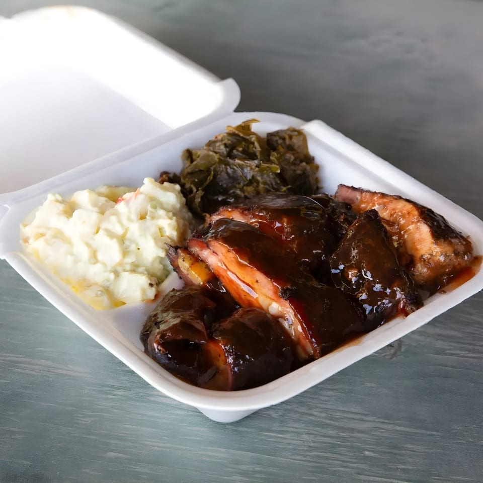 Mosito's BBQ ribs with dark sauce, potato salad, and collard greens in a white takeout container.