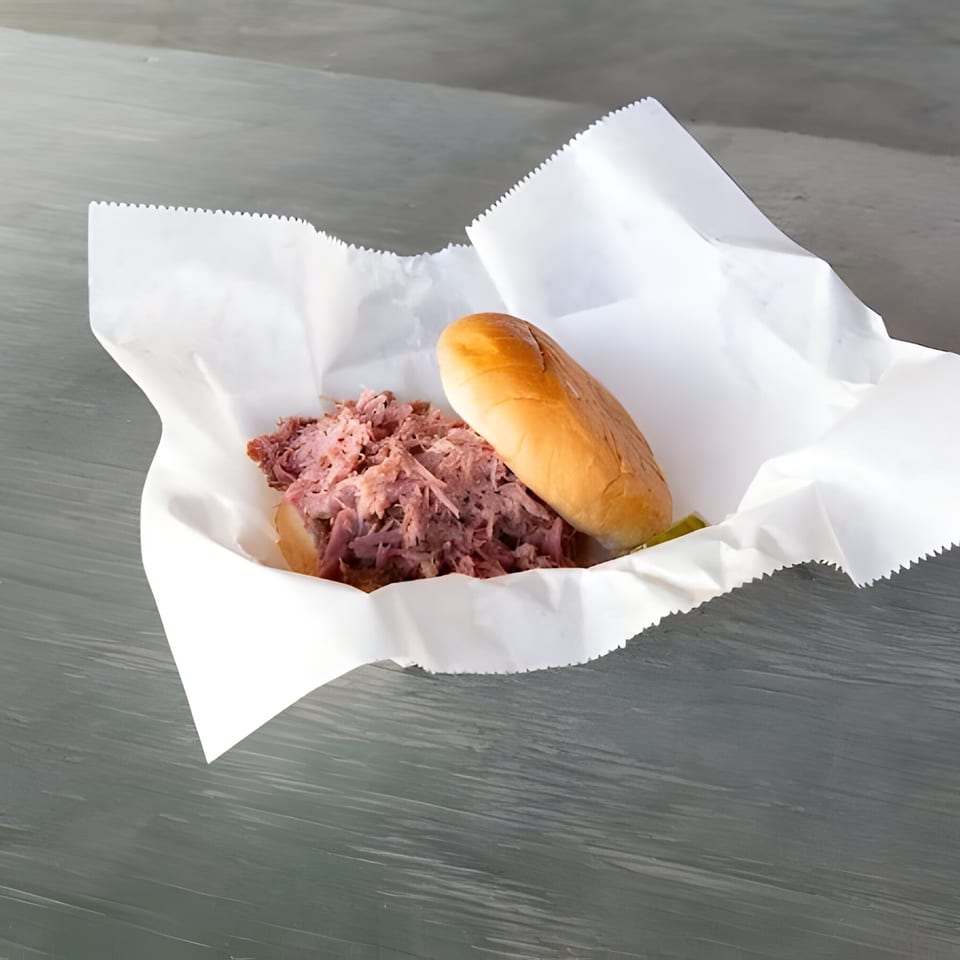 Mosito's BBQ pulled pork sandwich with a bun and pickles, served in white paper.