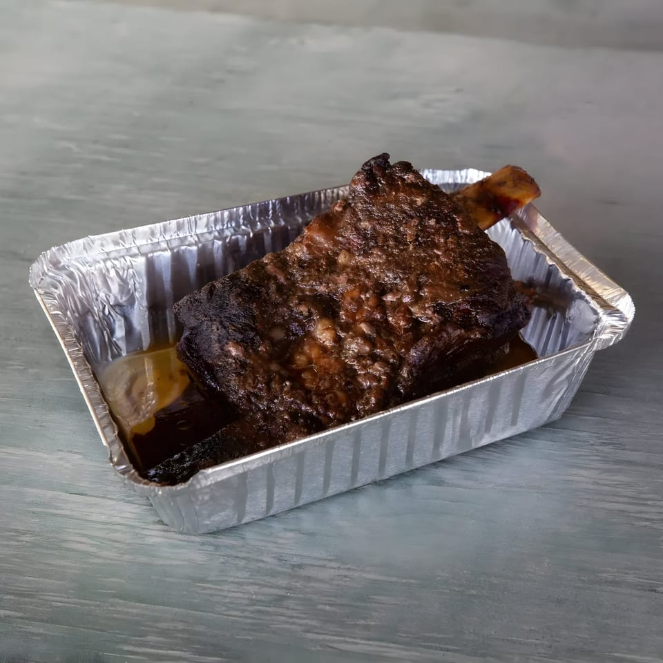 A large, dark, saucy BBQ beef rib in a silver foil container on a gray surface.