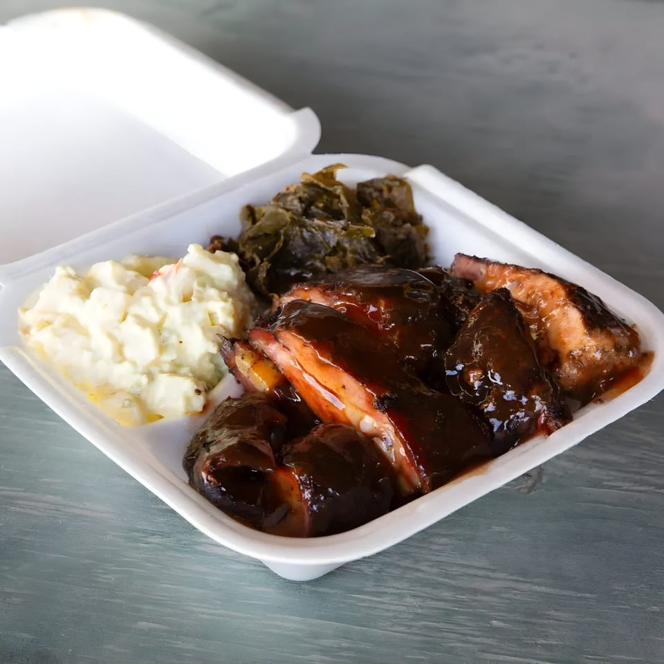 Mosito's BBQ ribs with dark sauce, potato salad, and collard greens in a white takeout container.
